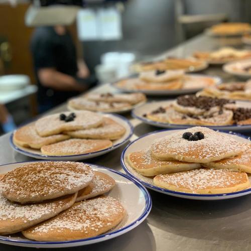 Stacks of pancakes dusted with powdered sugar and topped with blueberries, served on white plates in a busy kitchen.