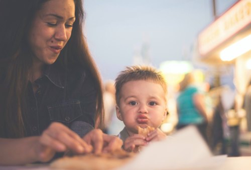A woman and a young boy enjoy food together at a lively outdoor event, with colorful lights in the background.