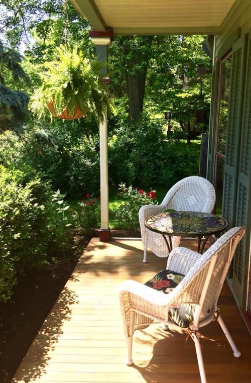 A cozy porch with two wicker chairs and a small table, surrounded by lush greenery and colorful flowers.