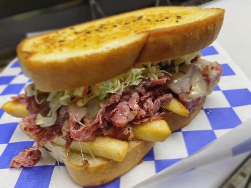 A hearty sandwich with shredded meat, melted cheese, fries, and coleslaw, served on toasted bread with a blue checkered wrapper.