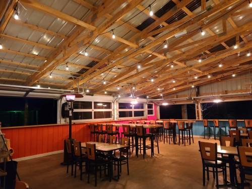 A cozy outdoor dining area with wooden tables, chairs, and string lights hanging from a wooden ceiling.