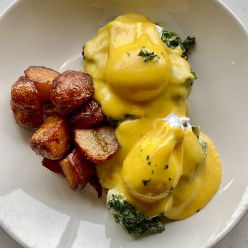 A plate of eggs Benedict with spinach and hollandaise sauce, served with crispy roasted potatoes on the side.