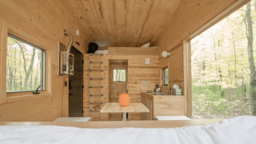 Cozy wooden interior of a tiny house with a kitchen, dining table, and large windows overlooking a forest.