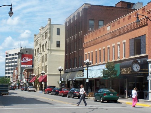 A bustling downtown street with shops, cars, and pedestrians under a blue sky with scattered clouds.