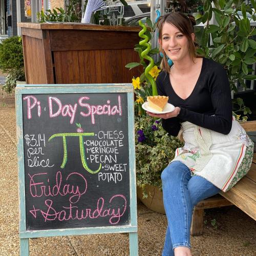 A woman in an apron holds a slice of pie next to a chalkboard sign advertising Pi Day specials.
