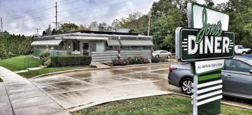 A retro-style diner with a green and silver exterior, surrounded by greenery and parked cars.