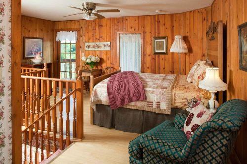 Cozy bedroom with wooden walls, a quilted bed, floral decor, and a staircase leading to a bright, inviting space.