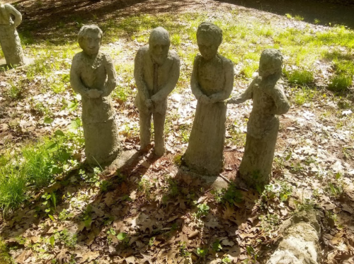 Four weathered stone sculptures of people standing in a grassy area, surrounded by fallen leaves and dappled sunlight.