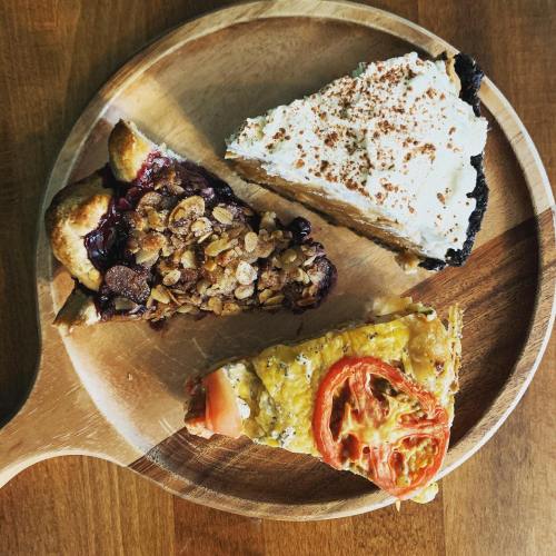 Three slices of pie on a wooden platter: berry crumble, cream pie, and savory quiche with tomatoes.
