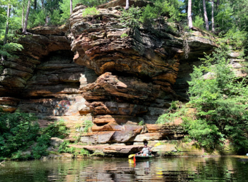 A person kayaking in a calm river surrounded by tall, rocky cliffs and lush green trees.