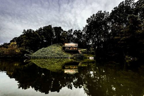 A modern house on a hill by a calm lake, surrounded by trees, reflecting in the water under a cloudy sky.