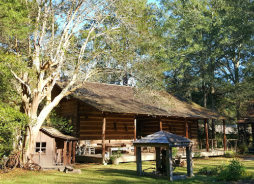 A rustic log cabin surrounded by trees, with a small shed and gazebo in a sunny outdoor setting.