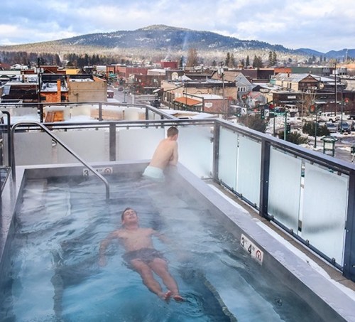 Two men relax in a rooftop hot tub with a scenic view of a town and mountains in the background.