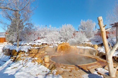 A steaming hot spring surrounded by snow-covered trees and rocks under a clear blue sky.
