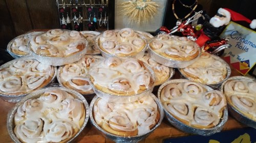 A display of multiple trays of cinnamon rolls, each topped with white icing, arranged on a table.