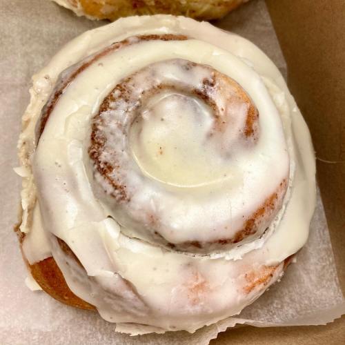 A freshly baked cinnamon roll topped with creamy white icing, resting on parchment paper.