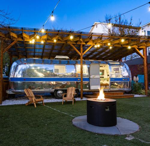 A shiny Airstream trailer under a wooden shelter, with string lights and a fire pit in a cozy outdoor setting.