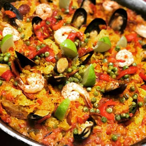 A vibrant paella featuring shrimp, mussels, bell peppers, peas, and lime, served in a large pan.