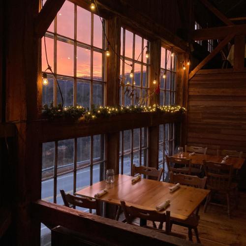 Cozy restaurant interior with wooden tables, warm lighting, and a sunset view through large windows.