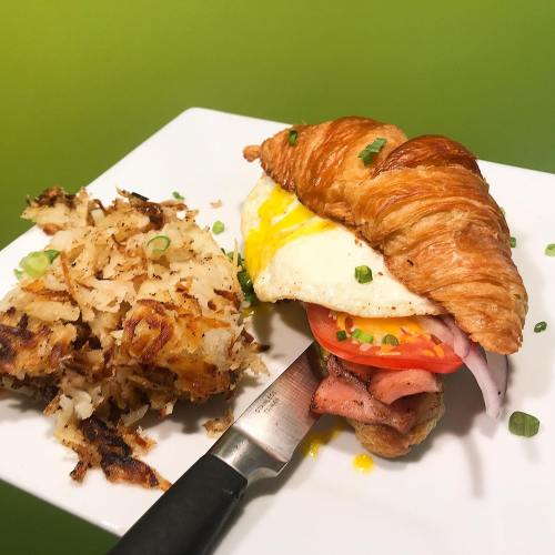 A croissant sandwich with ham, egg, and tomato, served with crispy hash browns on a white plate.