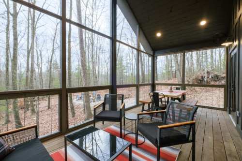 A screened porch with black chairs, a glass table, and a view of trees and a wooded area outside.