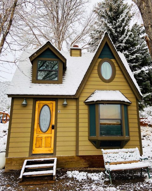 A charming, snow-covered cottage with a wooden door and green accents, surrounded by trees in a winter landscape.