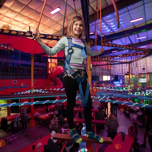 A young girl smiles while balancing on a high ropes course indoors, wearing a safety harness and colorful socks.