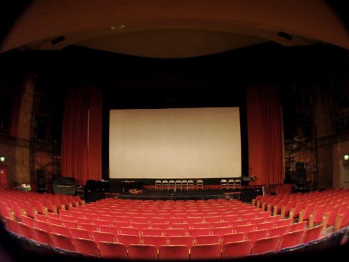 A wide view of a theater stage with a large blank screen and red seats arranged in rows.