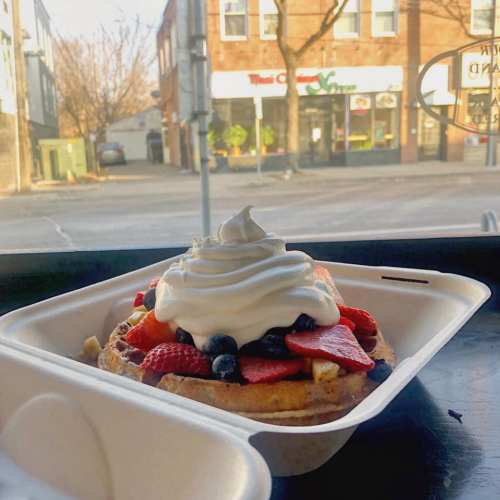A waffle topped with fresh strawberries, blueberries, and whipped cream, with a street view in the background.