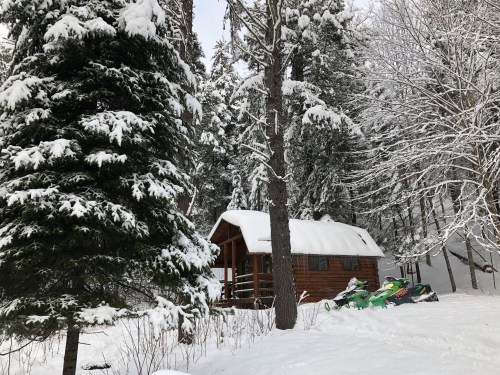 A cozy wooden cabin surrounded by snow-covered trees and snowmobiles in a winter landscape.