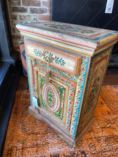 A colorful, vintage wooden cabinet with intricate floral designs, sitting on an orange patterned rug.
