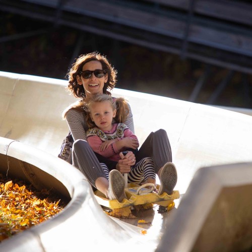 A woman and a child enjoy a ride on a luge track, surrounded by autumn leaves and sunlight.