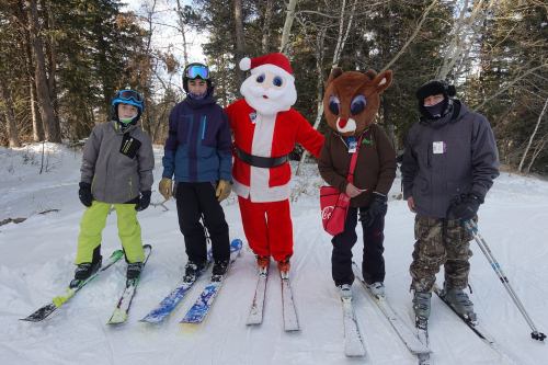 Four skiers pose on a snowy slope, including one in a Santa costume and another in a reindeer costume.