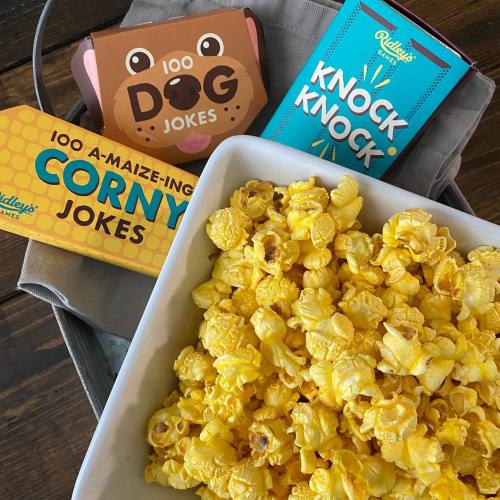 A bowl of yellow popcorn surrounded by joke card boxes: "100 Dog Jokes," "Knock Knock," and "100 A-Maize-Ing Corny Jokes."