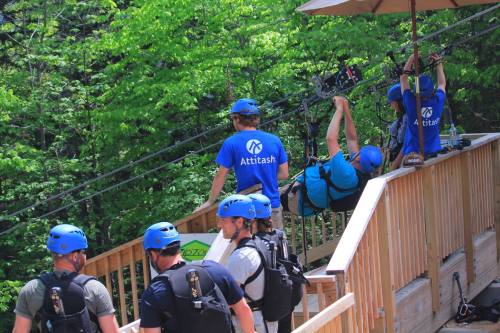 A group of people in blue helmets and shirts prepare for ziplining at a forested location.