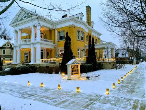 A large yellow mansion with white columns, decorated with lights, surrounded by snow and glowing lanterns along the path.