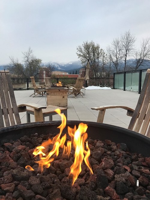 A cozy outdoor space with a fire pit, wooden chairs, and a view of mountains under a cloudy sky.
