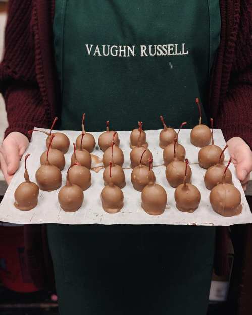 A person in an apron holds a tray of chocolate-covered cherries with stems, arranged neatly on parchment paper.