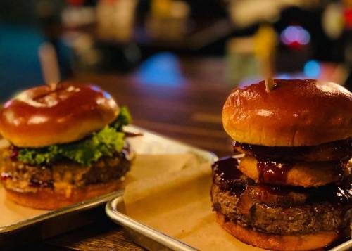 Two gourmet burgers on a wooden table: one with lettuce and the other stacked with onion rings and barbecue sauce.