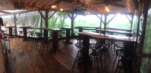 A rustic outdoor dining area with wooden tables and black chairs, surrounded by greenery and string lights.