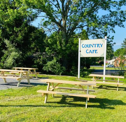 A sign for "Country Cafe" next to wooden picnic tables in a grassy area surrounded by trees.