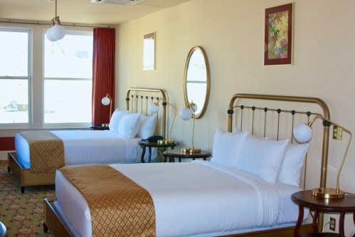 A bright hotel room featuring two beds with gold frames, a round mirror, and large windows with natural light.