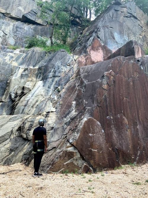 A person in climbing gear stands at the base of a large, rocky cliff, looking up at the steep rock face.