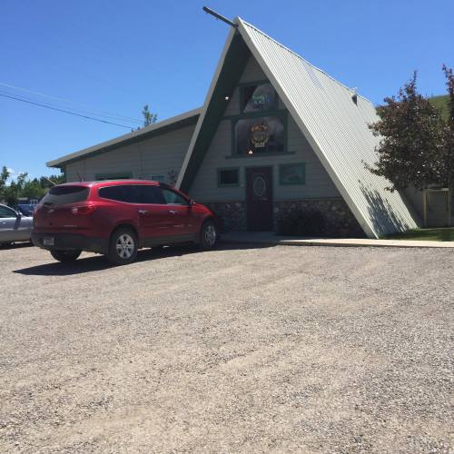 A red SUV parked in front of a triangular-roofed building on a sunny day, surrounded by gravel and greenery.