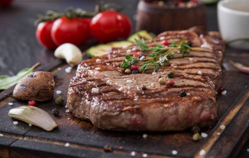A perfectly grilled steak garnished with herbs, surrounded by fresh tomatoes, garlic, and spices on a wooden board.