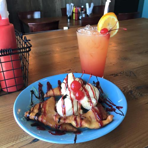 A blue plate with fried dessert topped with ice cream, chocolate sauce, and cherries, alongside a colorful drink with a slice of orange.