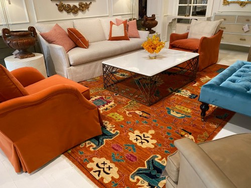 A stylish living room featuring a white sofa, orange chairs, a glass coffee table, and a colorful patterned rug.
