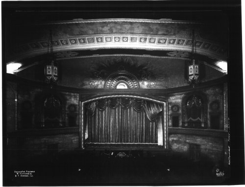 A vintage theater interior featuring an ornate stage with a curtain, decorative ceiling, and elegant lighting fixtures.