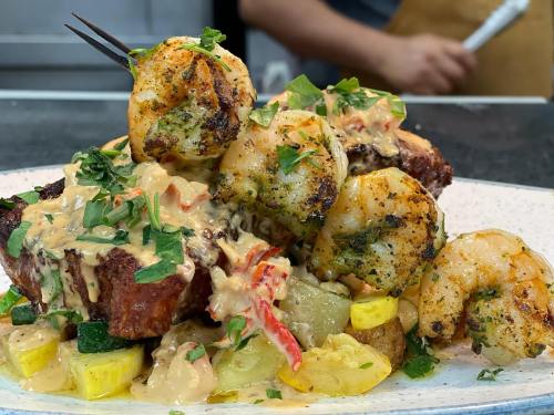 A plate of grilled shrimp atop seasoned vegetables, drizzled with a creamy sauce and garnished with fresh herbs.