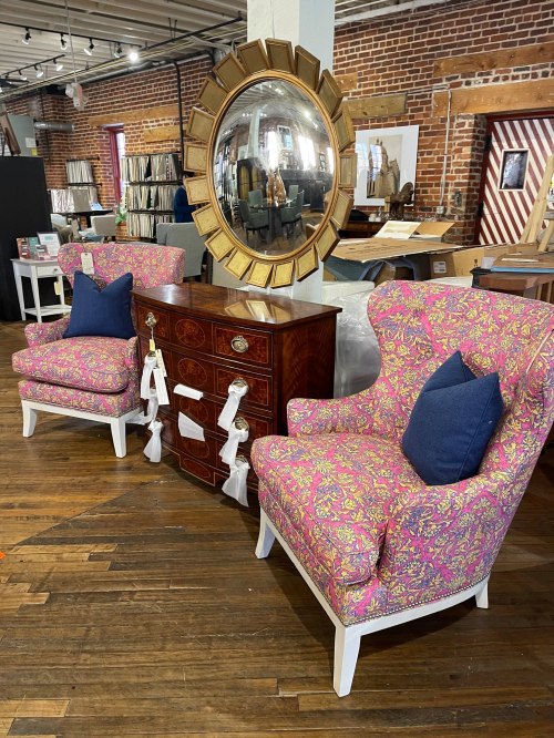 Two vibrant, floral-patterned chairs with blue pillows flanking a wooden dresser and a large round mirror in a stylish interior.